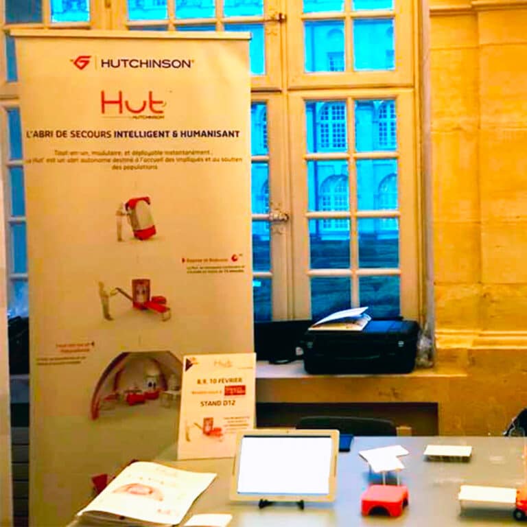 Signalétique grand format Hut’ by Hutchinson présentée sur un stand d’exposition, illustrant un abri de secours intelligent et humanisant développé par le groupe Hutchinson.